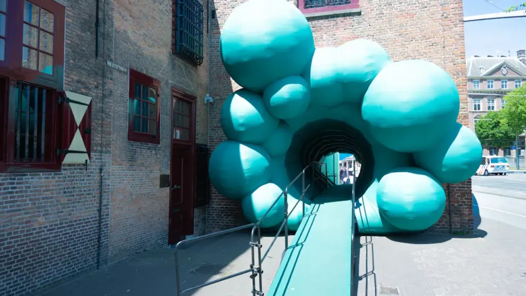 Türkisfarbene, begehbare Skulptur mit Rampe vor einem historischen Backsteingebäude im Freien.