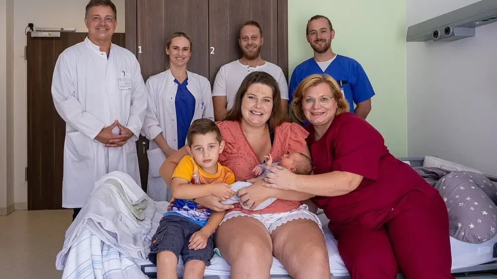 Eine junge Mutter mit ihrem Neugeborenen und ihrem Sohn, umgeben von medizinischem Fachpersonal im Krankenhaus.