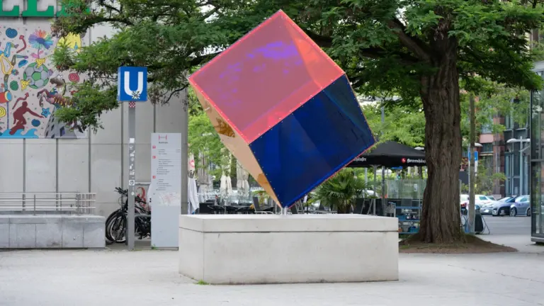 Eine moderne, schräg stehende Skulptur in Würfelform mit pinken und blauen Flächen.