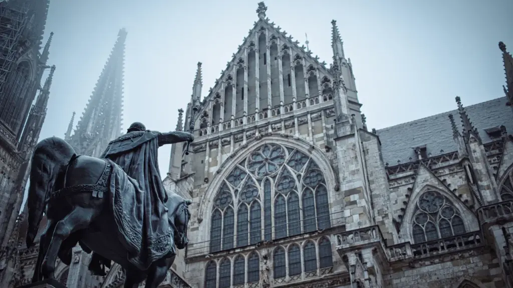 Historische Reiterstatue vor dem gotischen Regensburger Dom bei bewölktem Himmel.