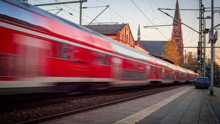 Ein vorbeifahrender roter Regionalzug vor der Kulisse einer deutschen Stadt bei Sonnenuntergang.