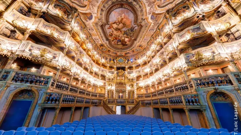Prunkvoller barocker Opernsaal mit goldenen Verzierungen, Deckengemälde und vielen blauen, leeren Sitzreihen im Vordergrund.
