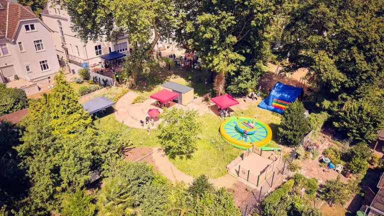 Vogelperspektive eines sonnigen Parks mit Hüpfburgen, Pavillons und spielenden Menschen im Grünen.