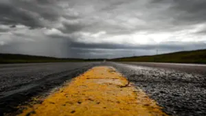 Nasse Landstraße mit gelber Fahrbahnmarkierung unter einem drohenden, dunklen Wolkenhimmel vor dem Unwetter.