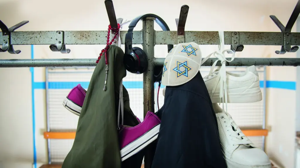 Eine Garderobe mit Kippa, Kopfhörern, roter Gebetskette, Jacken und verschiedenen Sneakern im Innenbereich.