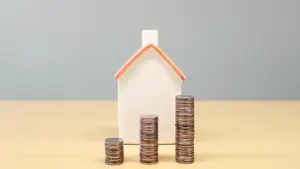 Ein kleines Hausmodell neben aufgetürmten Geldmünzen auf einem Holztisch als Finanzierungskonzept.