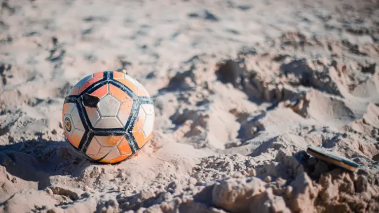 Ein abgenutzter Fußball liegt auf dem sandigen Boden eines Strandes bei hellem Tageslicht.