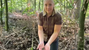 Eine blonde Frau im Wald hält einige braune Baumrindenstücke in ihren geöffneten Händen.