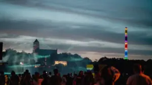 Menschenmenge bei Nacht vor einer beleuchteten Burg und einem leuchtenden, bunten Regenbogen-Schornstein.