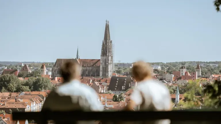 Blick auf den Regensburger Dom mit zwei Personen im unscharfen Vordergrund auf einer Bank.