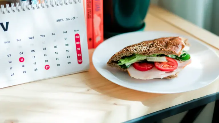 Ein Kalender und ein belegtes Sandwich auf einem Schreibtisch bei Sonnenlicht.