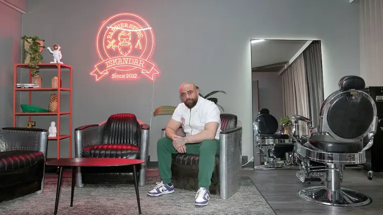 Ein bärtiger Mann sitzt in einem modernen Barbershop mit leuchtendem Neon-Logo an der Wand.