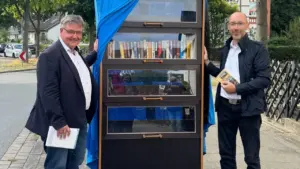 Zwei Männer stehen links und rechts neben einem hohen, dunkelbraunen Bücherschrank mit Glasfront im Freien.