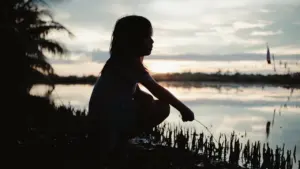 Die Silhouette eines jungen Mädchens hockt am Ufer eines Gewässers während eines friedlichen Sonnenuntergangs.