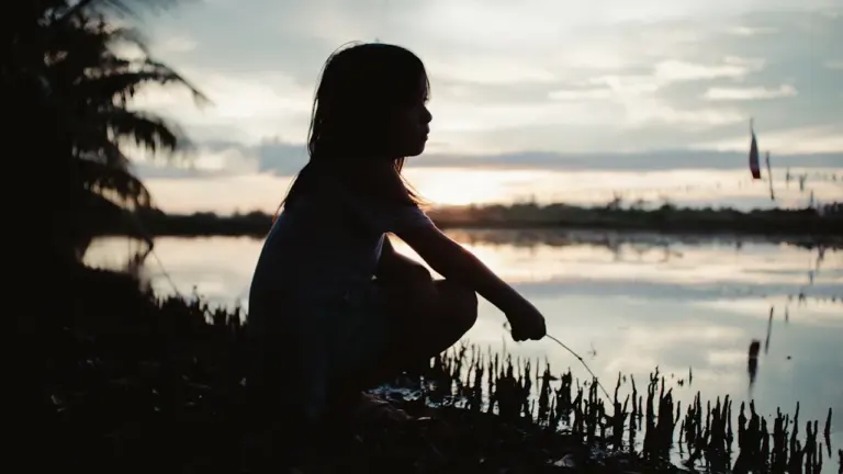 Die Silhouette eines jungen Mädchens hockt am Ufer eines Gewässers während eines friedlichen Sonnenuntergangs.